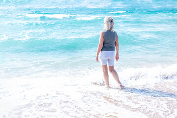 An elderly woman taking a walk by the sea. A happy lady in sportswear stands in the beautiful turquoise water and looks into the distance. Activity, peace, and freedom. Back view.