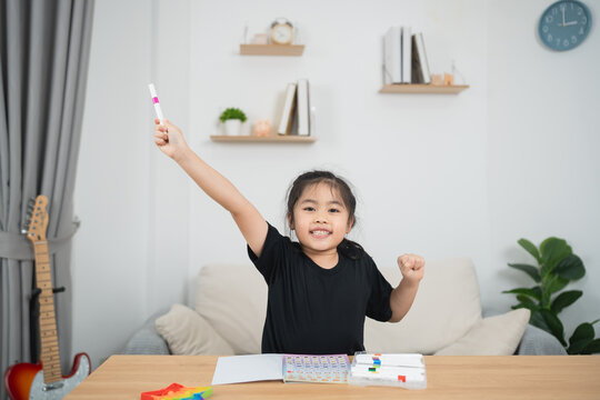 Cheerful girl in black shirt happily raising one hand holding a marker while sitting at a desk full of colorful art supplies and a cozy living room background