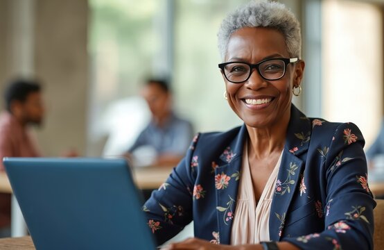 Smiling senior black businesswoman with gray hair wears glasses, uses laptop. She is happy and professional in an office setting. Her colleagues work in background. - Powered by Adobe
