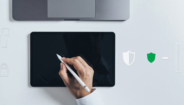 Hand using stylus on tablet device with laptop and security icons.