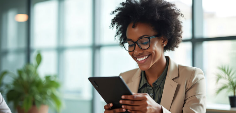 Smiling African American business woman uses digital tablet in bright modern office. Wears stylish glasses, happy to work online. Pro communicates, networks on social media, reviews important project