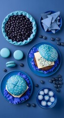 A vibrant blue-themed display of blueberries, macarons, cupcakes, and candies arranged on various plates and bowls for a visually appealing sweet treat presentation