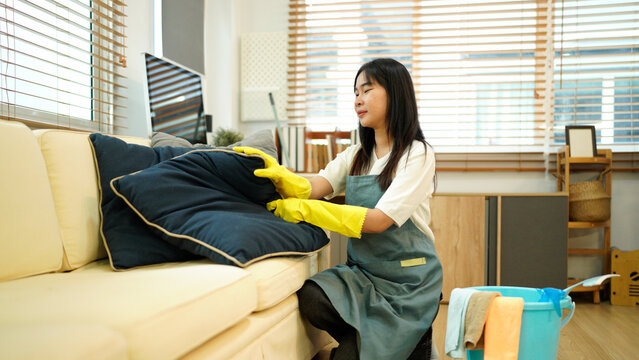 Housekeeper wearing yellow gloves is arranging pillows on a sofa in the living room, ensuring a tidy and comfortable environment for residents or guests - Powered by Adobe
