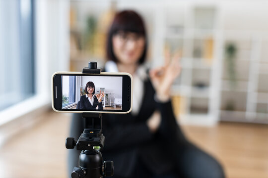 Caucasian adult woman in office wearing formal suit recording video blog. Office background, tripod setup visible. Business topic presentation representing professional, creative communication