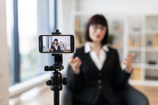 Caucasian woman recording video blog about business topics using smartphone camera on tripod. Professional indoor office setup emphasizes modern technology, content creation, and professionalism.