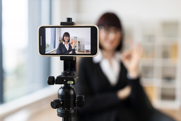 Caucasian woman wearing formal attire recording video blog using smartphone on tripod in modern office setting. Concept themes include business networking, educational content creation.
