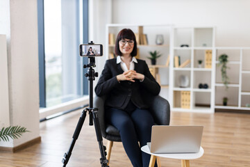 Caucasian adult woman using camera for recording business-related content in modern office environment. Focus on creating professional material remotely with digital tools.
