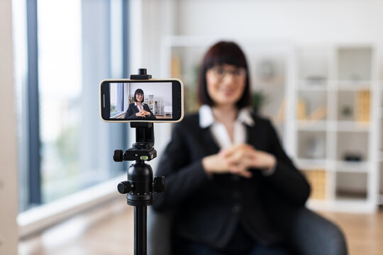Caucasian adult woman using camera for recording business-related content in modern office environment. Focus on creating professional material remotely with digital tools.