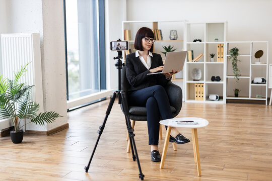 Caucasian businesswoman in modern office filming content on tripod camera holding laptop, delivering professional message. Bright interior with natural light highlights detailed setting