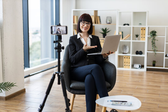 Young Caucasian woman in formal attire recording professional blog content on camera in spacious office environment, smiling while using laptop, showcasing modern working setting involving technology