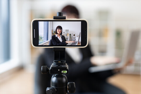 Caucasian woman in business attire recording herself on smartphone discussing business topics in office environment. Concept of video blogging, online content creation.