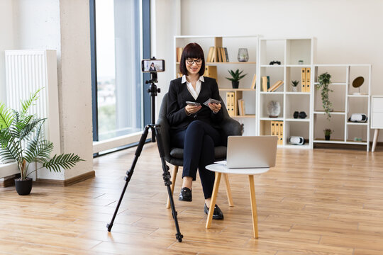 Caucasian woman in her thirties creating finance blog content holding cash dollars on camera. Depicts investment tips, blogging, and professional coaching in contemporary office with tripod