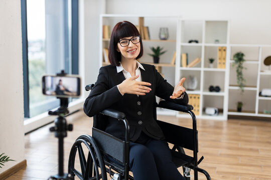 Caucasian woman wearing business attire sits in wheelchair recording video blog in office using smartphone. Professional setting conveys ideas of inclusivity accessibility business presentation