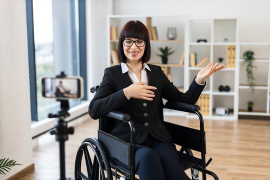 Caucasian woman wearing business attire sits in wheelchair recording video blog in office using smartphone. Professional setting conveys ideas of inclusivity accessibility business presentation