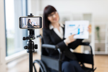 Caucasian woman wearing business attire in wheelchair records blog on smartphone tripod about business topics. Scene emphasizes professionalism, adaptability, modern business approach.