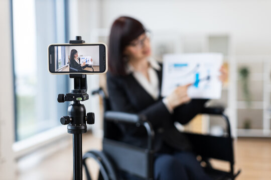 Caucasian woman in wheelchair blogs on camera about business using charts and graphs. Indoor setting with focus on professional blogging and business concepts.