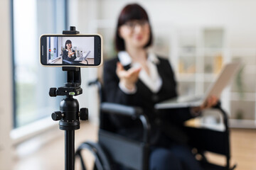 Caucasian woman in smart suit sitting in wheelchair in office during video blogging session about business practices using smartphone on tripod, promoting communication, inclusivity