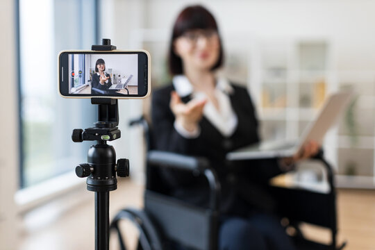 Caucasian woman in smart suit sitting in wheelchair in office during video blogging session about business practices using smartphone on tripod, promoting communication, inclusivity
