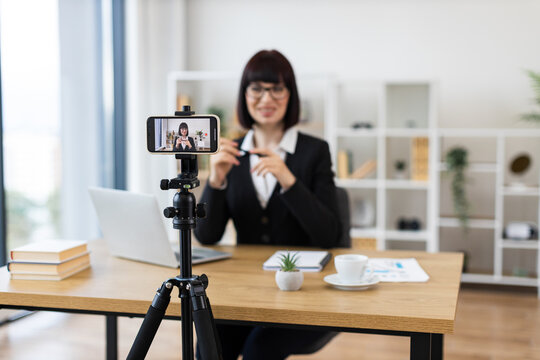Caucasian adult woman wearing black blazer in modern office using smartphone to record vlog showcasing business topics with creative desk setup that includes laptop and coffee cup.