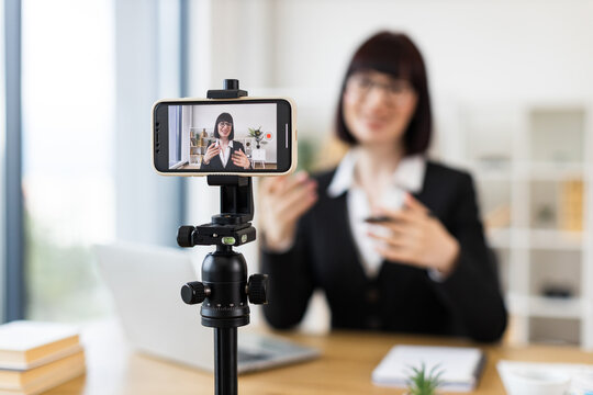 Caucasian woman in early 30s sitting at desk recording video blog about business-related topics.