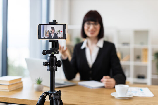 Caucasian woman wearing black suit recording video blog in office environment using smartphone on tripod