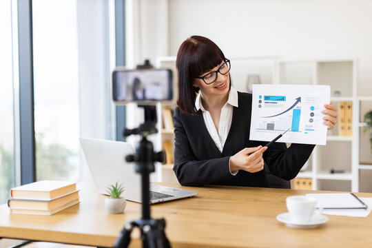 Caucasian adult woman with glasses at desk recording blog about business analysis and growth. Captures concepts like communication business trends remote work education and professional development.