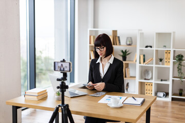 Caucasian woman, adult, wearing formal attire sitting at desk in office environment engaging in blog creation about investments, financial literacy, and income insights while holding dollar cash.