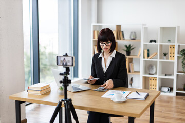 Caucasian woman, adult, wearing formal attire sitting at desk in office environment engaging in blog creation about investments, financial literacy, and income insights while holding dollar cash.