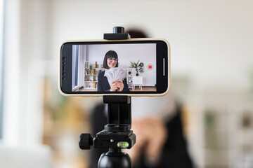 Caucasian woman mid 30s recording video blog about investments, financial literacy, income, holding cash, sitting at desk in modern office environment with smartphone on tripod for content creation