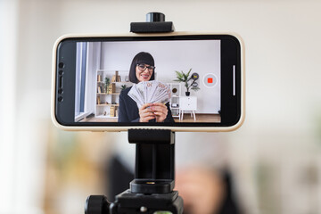 Caucasian woman mid 30s recording video blog about investments, financial literacy, income, holding cash, sitting at desk in modern office environment with smartphone on tripod for content creation