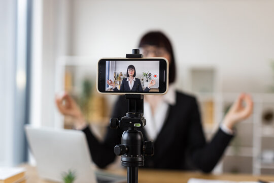 Caucasian woman in office environment recording meditation video at desk, combining work and relaxation. Professional appearance demonstrates how to balance workplace stress using mindfulness. - Powered by Adobe