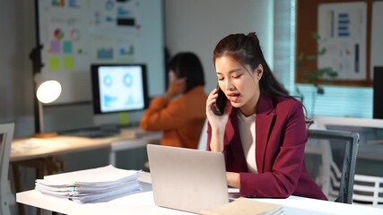 Young professional Asian businesswoman working late at night in a modern office, multitasking by talking on the phone and using her laptop to meet a deadline with her colleague - Powered by Adobe