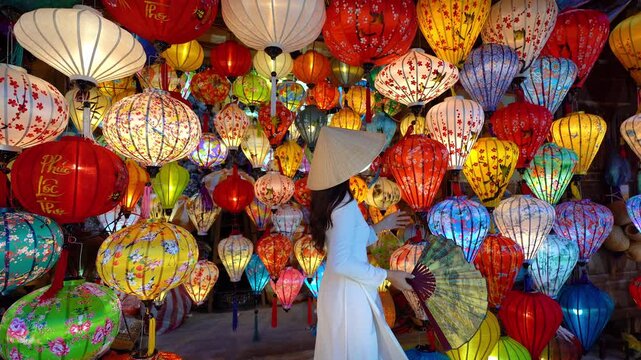 Beautiful Asian woman wearing traditional white ao dai dress with colorful paper lanterns in Hoi An, Vietnam.
