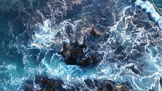Waves hit the dark rocks top view. Sea water splashes over the stones. Sea foam covers rocky beach. The blue ocean wave. Rocks break the sea waves. Water flows between the rocks. Foam moves.