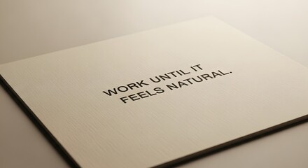 A close-up image of an open notebook with the motivational quote 'WORK UNTIL IT FEELS NATURAL' printed on a page, placed on a smooth surface with soft lighting