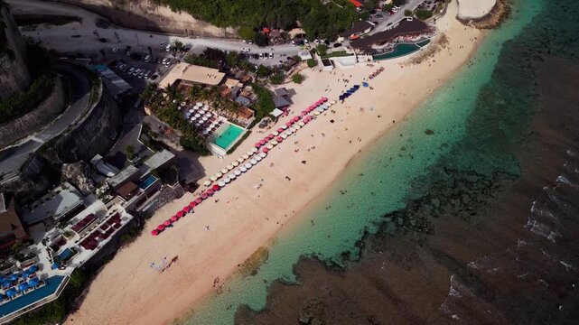 Aerial drone view of Melasti Beach Bali showing neat rows of umbrellas, turquoise sea, and a sunny tropical beach lounge surrounded by limestone cliffs and calm ocean waters on a bright island day.