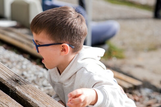 Excited young boy in a gray hoodie and blue glasses leaning over an outdoor wooden surface, engaged in play or observation
