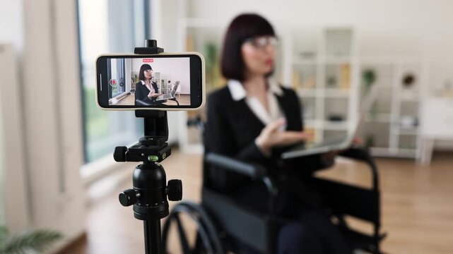 Caucasian woman in smart suit sitting in wheelchair in office during video blogging session about business practices using smartphone on tripod, promoting communication, inclusivity