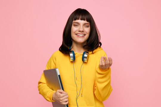Bright portrait of a cheerful student in a yellow hoodie, holding a laptop and headphones, against a soft pink backdrop, conveying focus and creative energy.