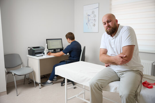 Man suffering from elbow pain during medical consultation in physiotherapy clinic. Patient holding arm in discomfort while doctor examines his shoulder. 