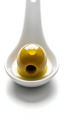 A close-up of a white ceramic spoon holding a green olive with a small hole, set against a plain white background, highlighting the smooth texture and glossy surface of the olive