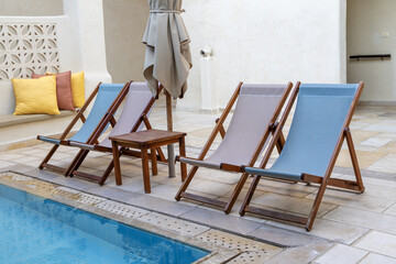 Poolside scene featuring three wooden lounge chairs with fabric cushions, a small wooden table, and...