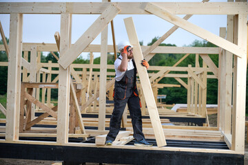 Construction worker installing wooden beam at house frame construction site. Carpenter in safety gear building timber structure for new home outdoors.
