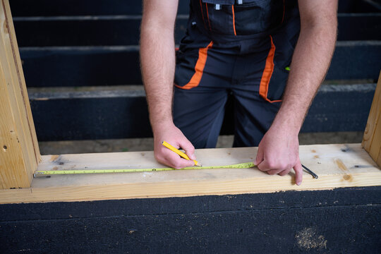 Close-up of construction worker marking wooden beam with pencil and tape measure at building site. Precision measurement during house frame construction.