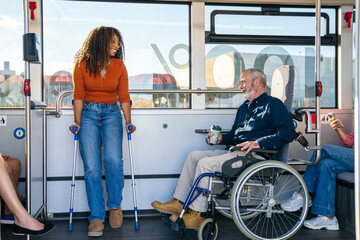 African woman with crutches and senior man in wheelchair smiling and interacting on a modern city bus © Koldo_Studio