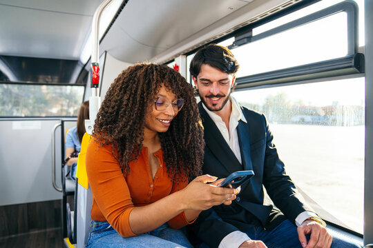 Diverse couple traveling on a public bus, smiling while browsing content on a smartphone, connecting and commuting in the city