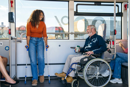 African woman with crutches and senior man in wheelchair smiling and interacting on a modern city bus - Powered by Adobe