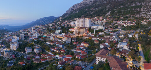 Twilight aerial panorama of Krujë showing the old bazaar, modern hotels, and mountain slopes under soft evening light.