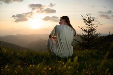 Woman sits peacefully on a grassy hill, gazing at the sunset as golden light bathes the landscape. Rolling mountains and a single evergreen tree create a tranquil atmosphere in the evening.