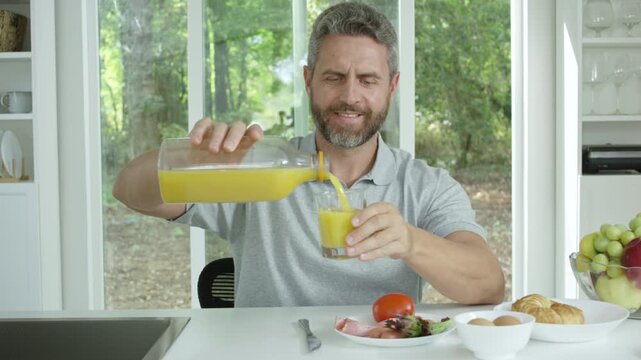 Man pouring orange juice at morning kitchen. Mature man pours fresh orange juice for breakfast. Middle aged nan eating breakfast in cozy kitchen.